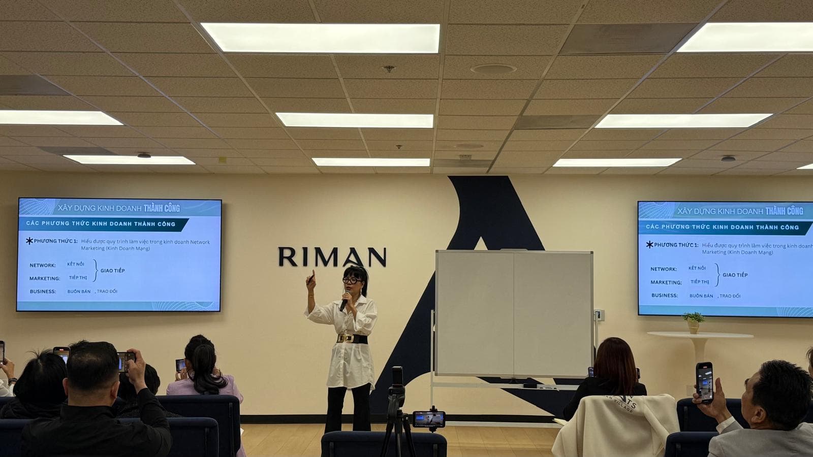 A presenter speaking into a microphone beside screens and a whiteboard as attendees watch.