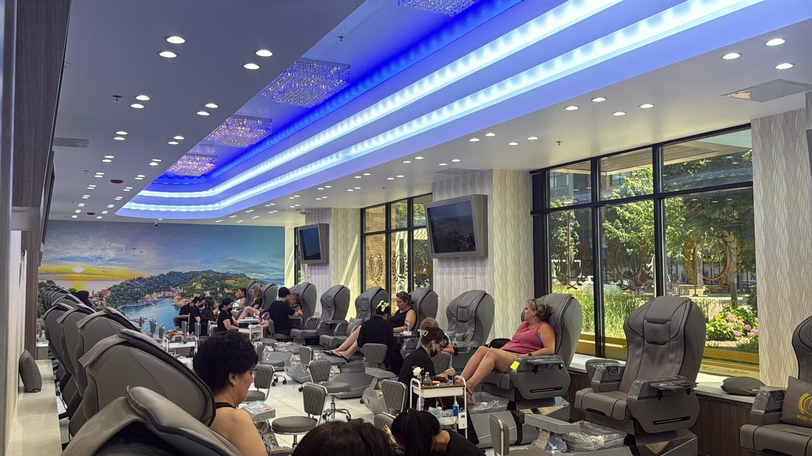 Clients and beauty professionals in a salon with rows of chairs and large windows.